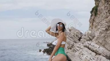 一位穿着绿色<strong>比基尼</strong>、太阳镜和大帽子的白色苗条深褐色头发的<strong>女士</strong>，在阳光明媚的岩石附近的海滩上摆姿势